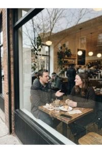 Two adults talking across a small cafe table beside a street-facing window.