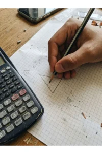 A hand sketching a geometric shape on graph paper beside a calculator.