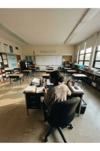 An adult working at a teacher's desk in a bright classroom.