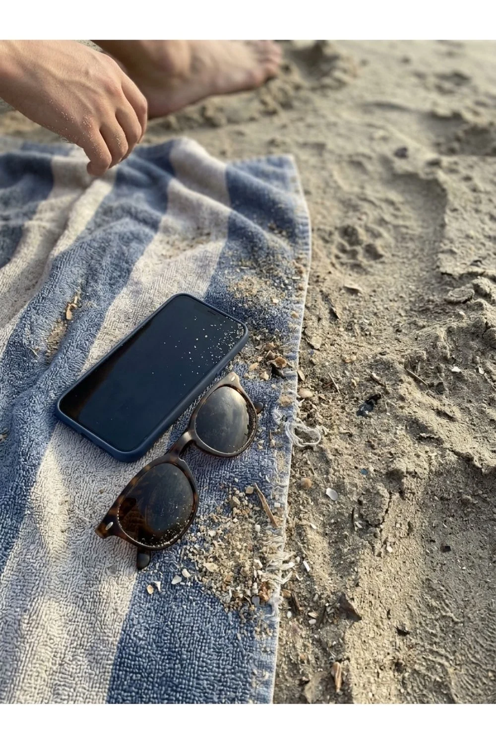 A beach towel and sunglasses on sand near the water.