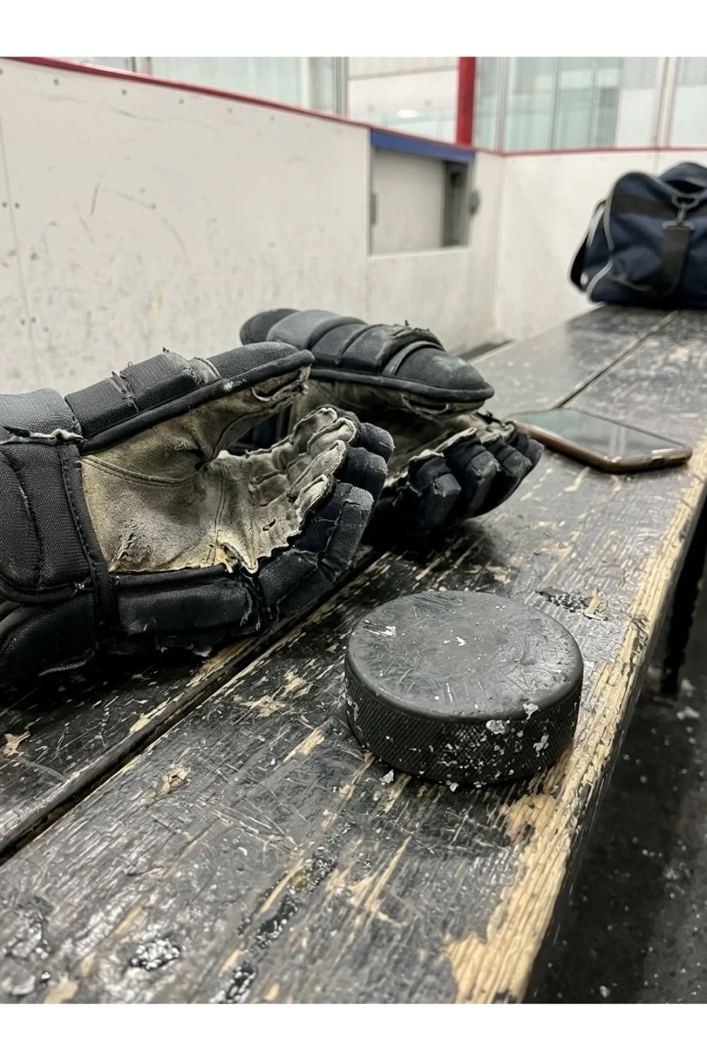 Used hockey gloves and a puck on a wooden bench beside a rink.