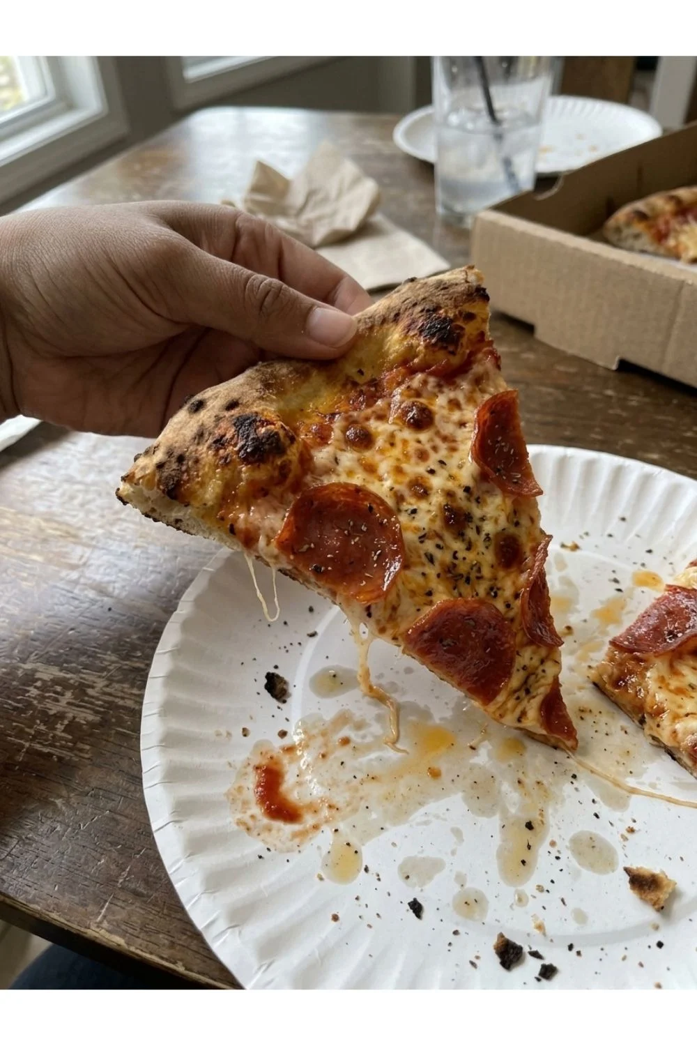A hand holding a slice of pizza over a used paper plate.