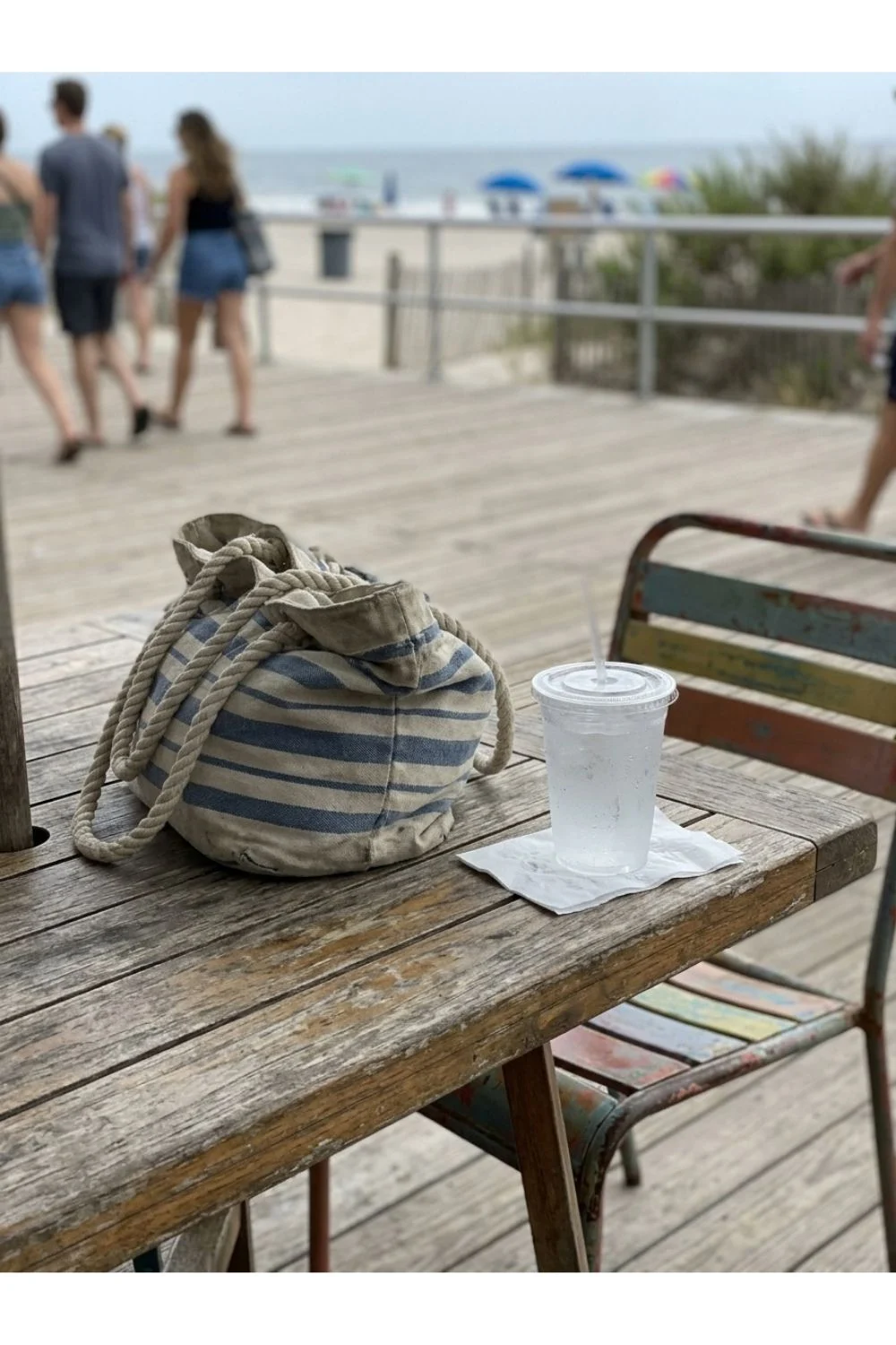 Beach bags and a drink on a picnic bench near a boardwalk.